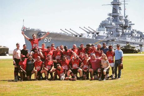 A rugby team in red jerseys poses for a group photo on grass in front of a large battleship.