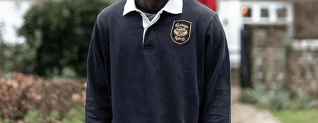 A person with long hair sits outdoors on a chair, wearing a navy and white striped shirt with a "USA Rugby" logo. Trees and sunlight form the background scenery.