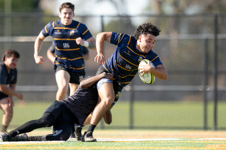 A rugby player holds the ball while being tackled, with teammates in the background on a grassy field.