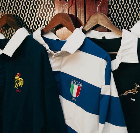 Collection of vintage rugby shirts with country emblems hanging on wooden hangers against a wooden backdrop.