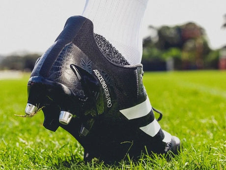 Close-up of a person wearing black and white Adidas Adizero RS7 Pro cleats standing on a green field.