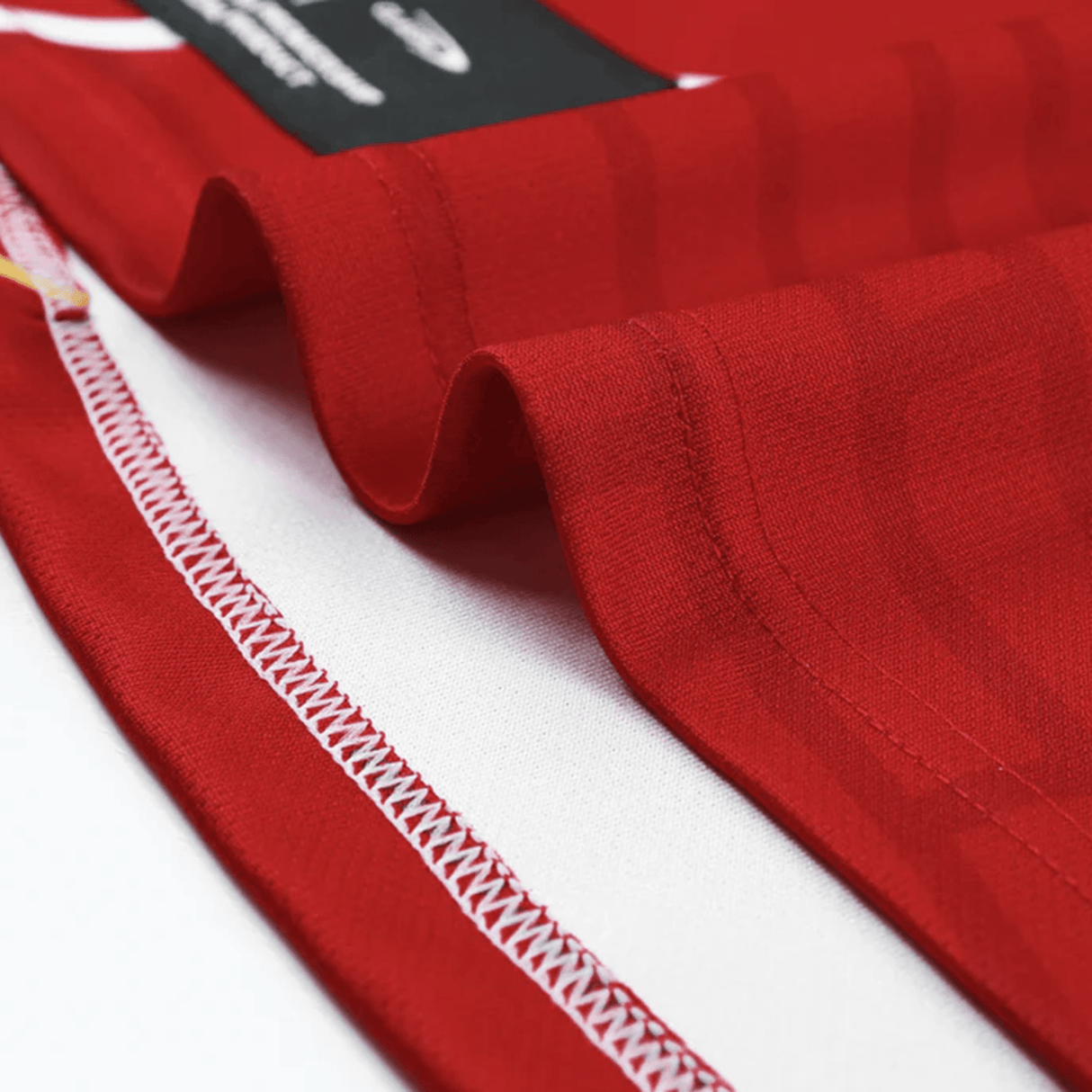 Close-up of red fabric with white stitching along the edge. The fabric is slightly folded, revealing a small part of a black label. This detail is reminiscent of the Dolphins NRL Heritage Jersey by Classic Sportswear, exemplifying classic sportswear style.