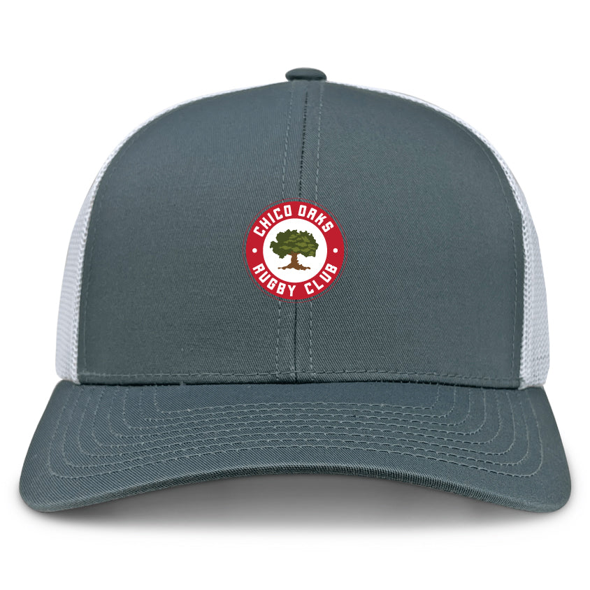 The Chico Oaks Retro Trucker Cap by EMB Alpha Broder is a gray and white trucker cap featuring the "Chico Oaks Rugby Club" logo with an embroidered oak tree in a red circle and snapback closure for adjustable comfort.