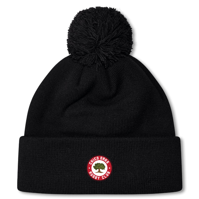 The Chico Oaks Pom Pom Beanie by EMB Canterbury is a black beanie featuring a red and white "Chico Oaks Rugby Club" patch with an oak tree, topped with a pom-pom and fleece-lined for warmth, perfect for any Canterbury rugby enthusiast.