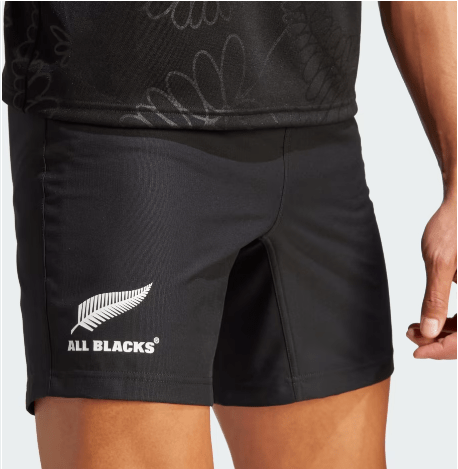 Close-up of a person wearing All Blacks Home Short by adidas in black, featuring the iconic silver fern emblem on the left leg. Made from recycled polyester and incorporating AEROREADY technology for comfort, these shorts are branded by adidas. The person’s hand is partially visible.