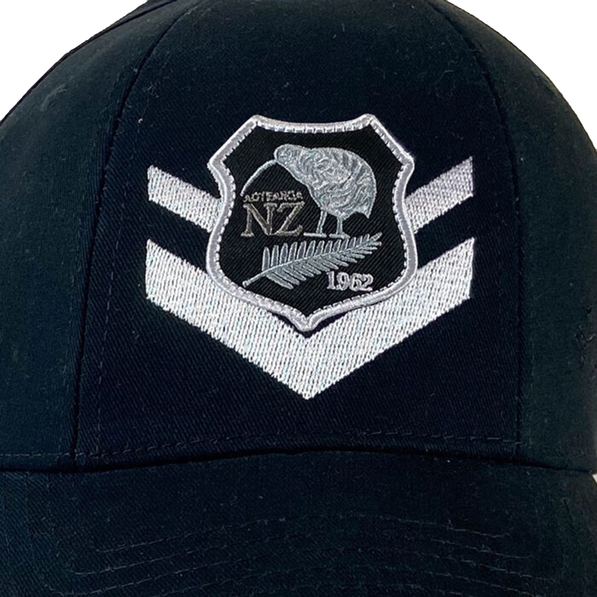 The New Zealand 1962 Rugby League Cap by Ellis Rugby, a black cap with a silver embroidered emblem showcasing a kiwi, "NZ," and "1962" on the front, commemorates Kiwi sports history. It also features two white chevrons stitched beneath the emblem.