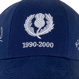 The Ellis Rugby Doddie Weir Scotland Cap features navy blue fabric with an embroidered thistle emblem and the years "1990-2000" in silver stitching, evoking Doddie Weir's glory days.