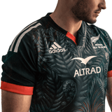 A person wearing the Māori All Blacks Home Supporters Jersey by adidas, designed in black and crafted from recycled polyester. This stylish adidas jersey showcases the Altrad logo and features moisture-wicking fabric for ultimate comfort.