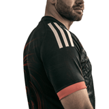 The man wearing the adidas Māori All Blacks Home Supporters Jersey, featuring a black design with white stripes and red trim, stands sideways against a white background. The jersey is made from recycled polyester and includes moisture-wicking fabric for top-notch performance.