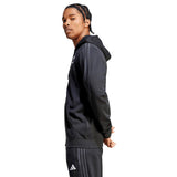 A person in an All Blacks Supporter Full-Zip Hoodie by adidas, crafted from French terry fabric, and track pants featuring white stripes is standing sideways with their hands behind their back against a plain white background.