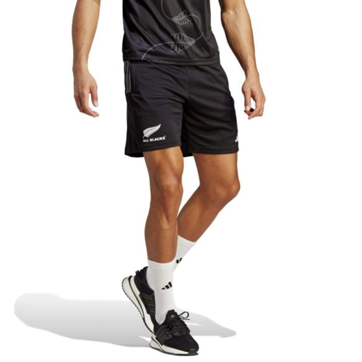 A man wearing a black sports outfit, including a "New Zealand All Blacks" t-shirt and adidas All Blacks Gym Short featuring moisture-wicking AEROREADY technology, white socks, and black running shoes, standing with one leg slightly forward.