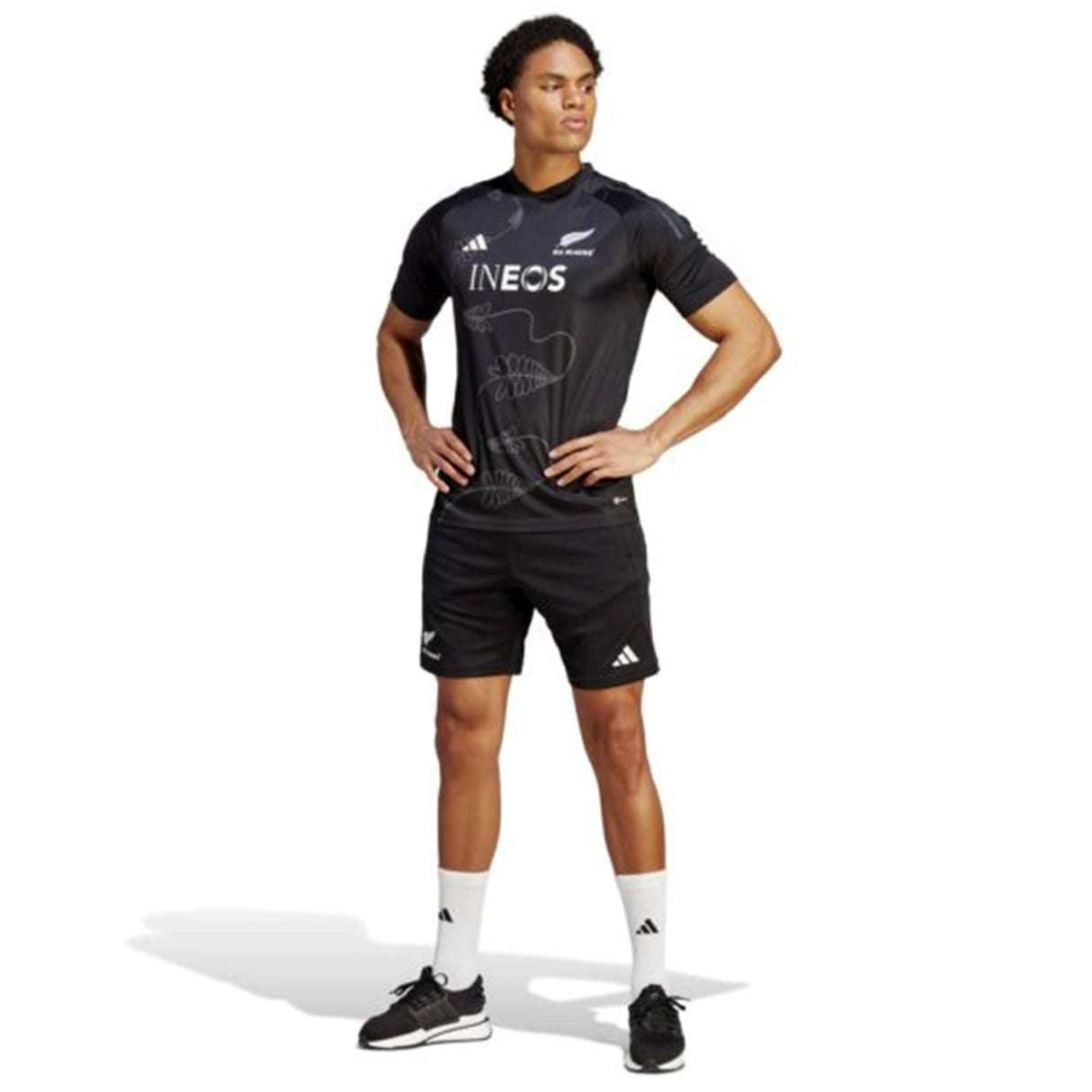 A man stands with his hands on his hips, wearing a black sports uniform with "INEOS" branding, white socks, and black sneakers. His All Blacks Gym Shorts by adidas feature moisture-wicking AEROREADY technology, reminiscent of the New Zealand All Blacks' iconic gear.