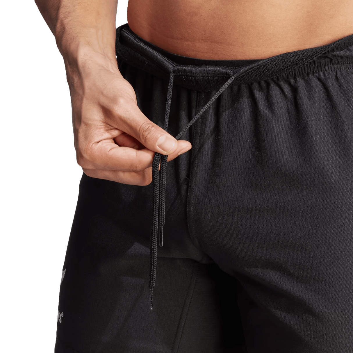 Close-up of a person's hand tightening the drawstring on a pair of black All Blacks Home Shorts by adidas made from recycled polyester, with part of their bare abdomen visible.