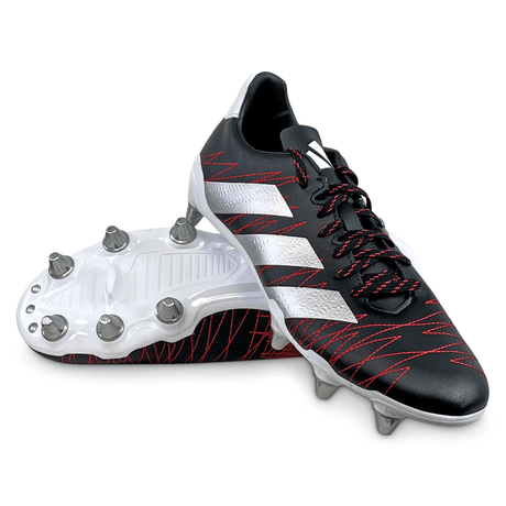 A pair of adidas Kakari SG rugby boots in Core Black/White/Better Scarlet, featuring a design with white stripes and red accents. Crafted from recycled materials, one boot stands upright while the other lies on its side displaying metal studs. Ideal for soft ground, these adidas boots are engineered to enhance on-field performance.