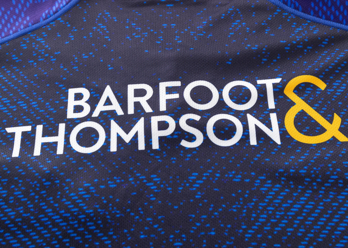 Close-up of the Blues Super Rugby 25/26 Replica Home Jersey by Classic Sportswear, featuring "BARFOOT & THOMPSON" text in white and yellow on a dark blue patterned background.