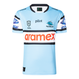 The Sharks NRL 25/26 Replica Home Jersey by Classic Sportswear features a light blue design with black and white striped sleeves, adorned with Pilot, Aramex, and Cronulla Sutherland Sharks logos. A must-have for any Sharks fan.