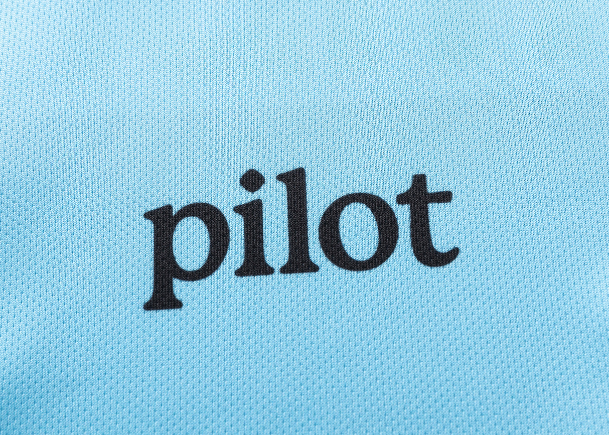 The word "pilot" is printed in black lowercase on a textured light blue fabric, reminiscent of the Sharks NRL 25/26 Replica Home Jersey by Classic Sportswear.