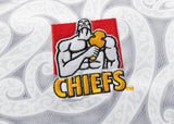 A muscular figure holding a hammer embroidered above "CHIEFS," set on a red square, adorns patterned fabric inspired by the Gallagher Chiefs Super Rugby Replica Away Jersey 25/26 by Classic Sportswear.