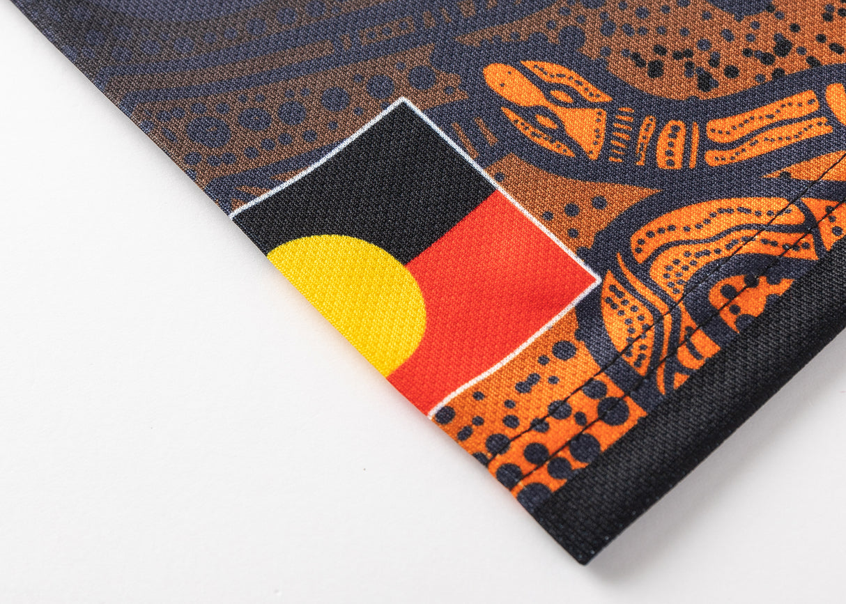 A fabric close-up shows a section of the Aboriginal flag in black, yellow, and red with intricate orange and black patterns, mirroring the design found in the NRL Indigenous All Stars 25/26 Replica Home Jersey by Classic Sportswear.