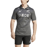 A person wearing a gray All Blacks Training Jersey by adidas featuring a French Fern design and the word "INEOS" on the chest, paired with black shorts, standing and looking slightly to the side.