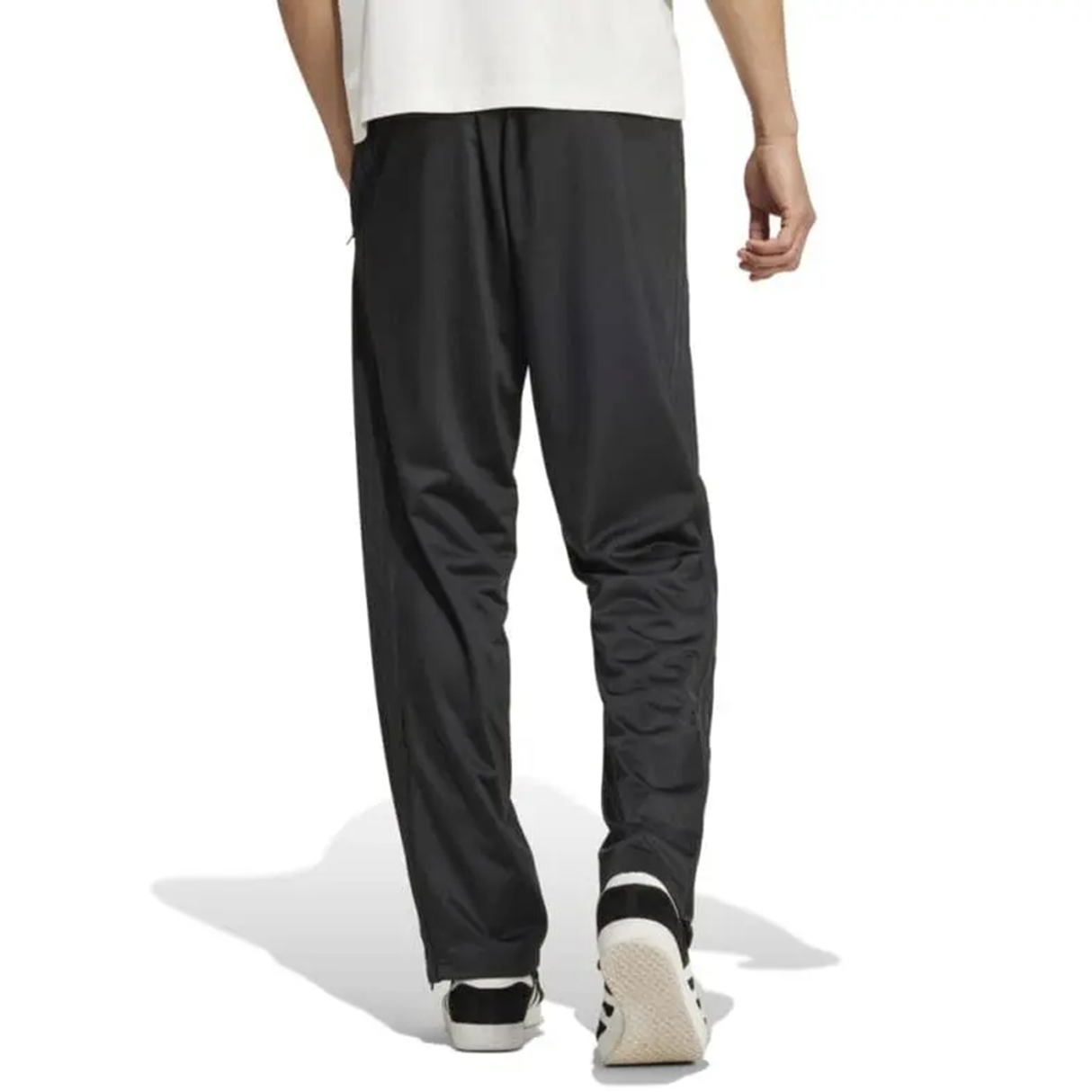 A person walking away on a white background, wearing a white shirt, Originals Limited Edition All Blacks Firebird Track Pants by adidas, and black and white sneakers.