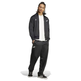 A person in Originals Limited Edition All Blacks Firebird Track Pants by adidas and a matching track jacket stands confidently against a plain white backdrop, complemented by a crisp white t-shirt and sleek black shoes.