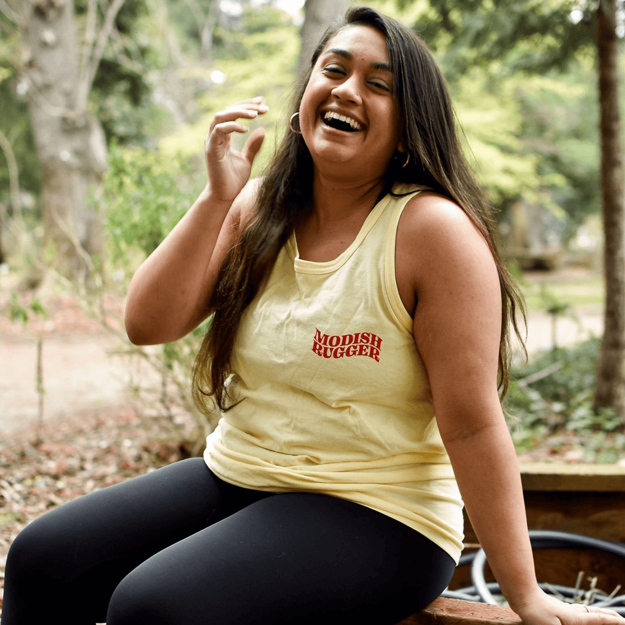 A person sits outdoors on a wooden bench, wearing a Modish Rugger Saturday's a Rugby Day Tank Top paired with black leggings, smiling and laughing amidst the trees.