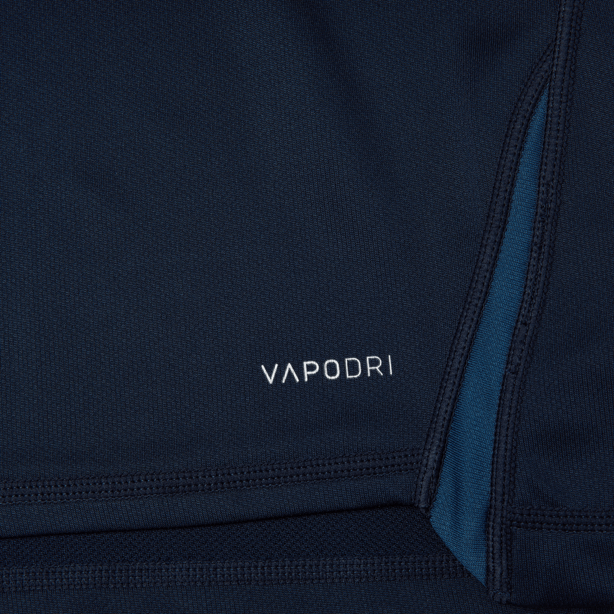 Close-up of a dark blue fabric with the word "VAPODRI" printed in white letters, accompanied by teal stitching detail, highlighting the Canterbury Vapodri Evader Jersey's advanced moisture-wicking technology.