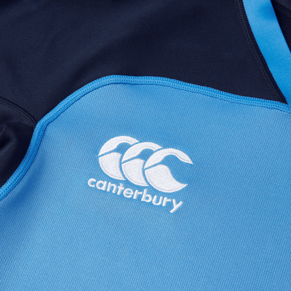 Close-up of a Canterbury Vapodri Evader Jersey with the Canterbury logo, featuring three interlocking Cs, embroidered in white on a blue fabric background. This jersey boasts moisture-wicking technology and a VapoDri finish to keep you comfortable and dry.