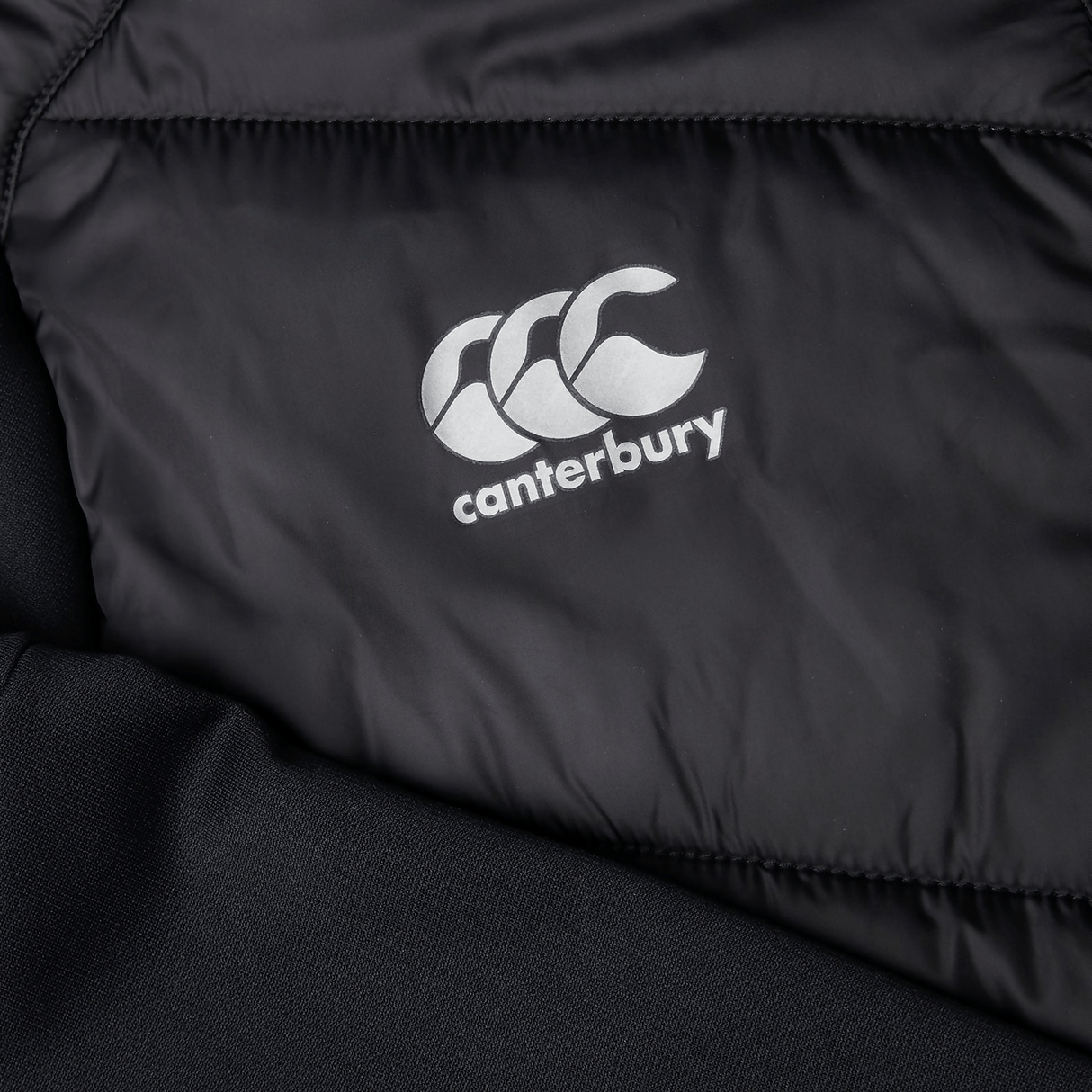 Close-up of the black Canterbury Hybrid Jacket V2 showcasing a white logo with three stylized C's, featuring ThermoReg technology and a water-resistant finish.