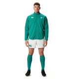 A man wearing a Canterbury Ireland 24 Anthem Jacket in green with water-resistant fabric technology, white shorts, green knee-high socks, and black cleats stands against a plain white background.