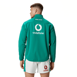 A person is wearing a green Ireland 24 Anthem Jacket by Canterbury and white shorts with brand logos. The back of the jacket features "vodafone" and "IRELAND" printed on it. This jacket boasts water-resistant fabric technology, making it both stylish and practical.