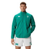 A person stands wearing the Ireland 24 Anthem Jacket by Canterbury, which features heat-pressed logos, along with white shorts adorned with green logos. They pose against a plain white background.