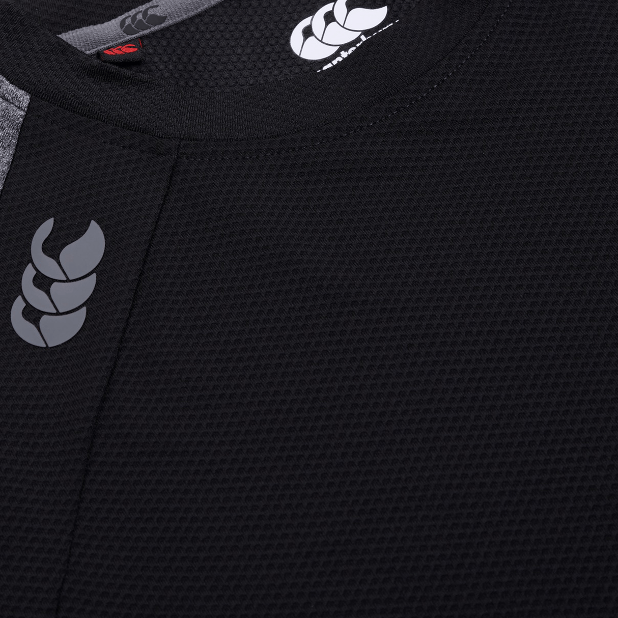 Close-up of a Women's Canterbury Elite Training Tee, highlighting its black textured sports jersey design featuring a gray shoulder logo. Crafted with Vapodri technology, its lightweight fabric promises optimal performance against a subtle gray background.