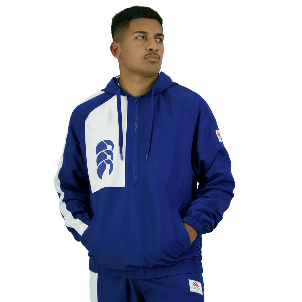 A man stands wearing a blue and white Legends Woven 1/4 Zip hooded tracksuit by Canterbury, hands in pockets, looking to the side against a white background. This sports casualwear piece embodies the relaxed yet dynamic style of the collection.