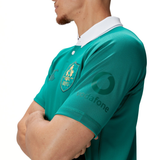 A person in a green Ireland 150th Anniversary Pro Home Jersey by Canterbury, featuring a white collar, stands with arms crossed. This jersey proudly displays the logo and sponsor name on the sleeve.