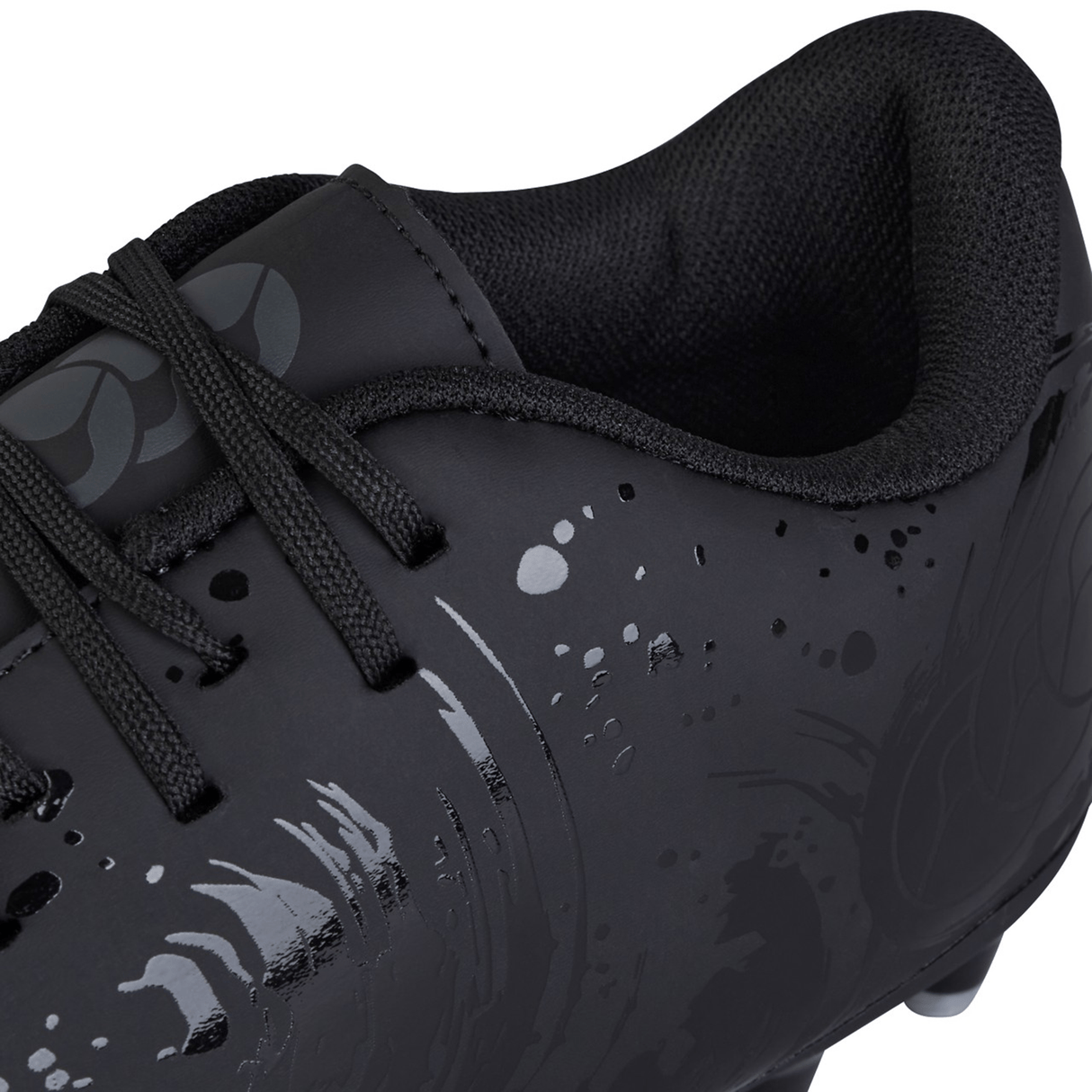 Close-up of a Canterbury Phoenix Genesis Team FG athletic shoe in black and silver metallic, featuring dark laces, a shiny abstract pattern on the synthetic leather forefoot and side area, and a cushioned, textured inner lining.