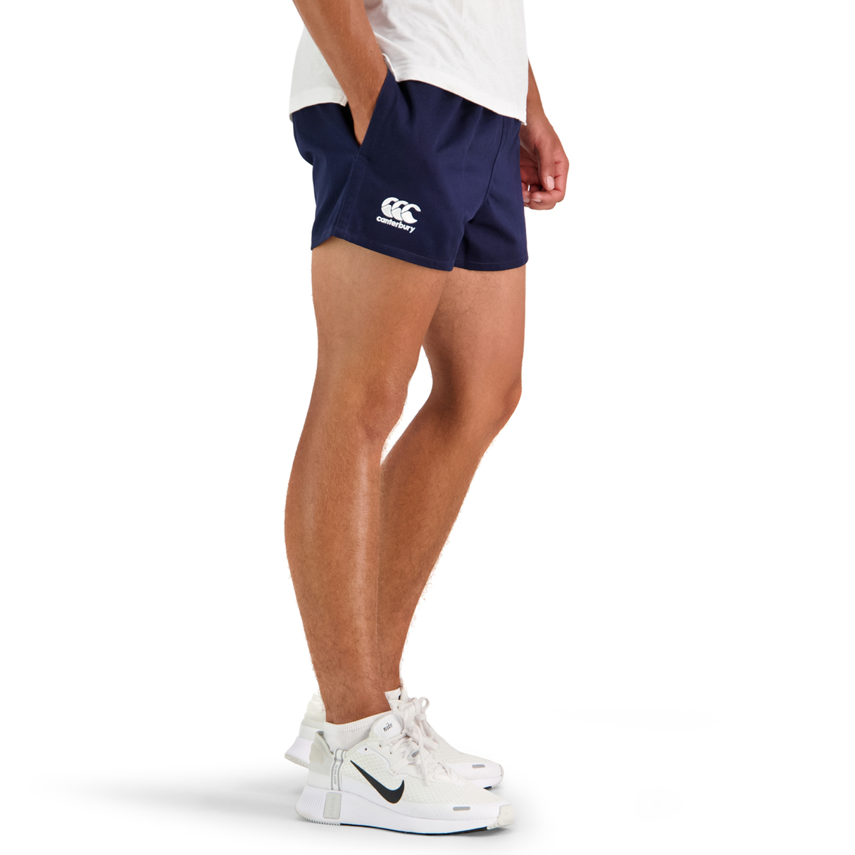 A person stands in a side profile wearing white sneakers, a white shirt, and navy Canterbury Classics Rugged Drill Shorts with a logo.