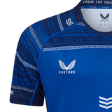 Leinster 24/25 Replica Home Jersey by Castore, a blue athletic shirt with patterned sleeves and neckline, prominently displays the Castore logo and text. This design features an athletic fit and proudly showcases the Irish Harp graphic, embodying the spirit of a Leinster Rugby jersey.