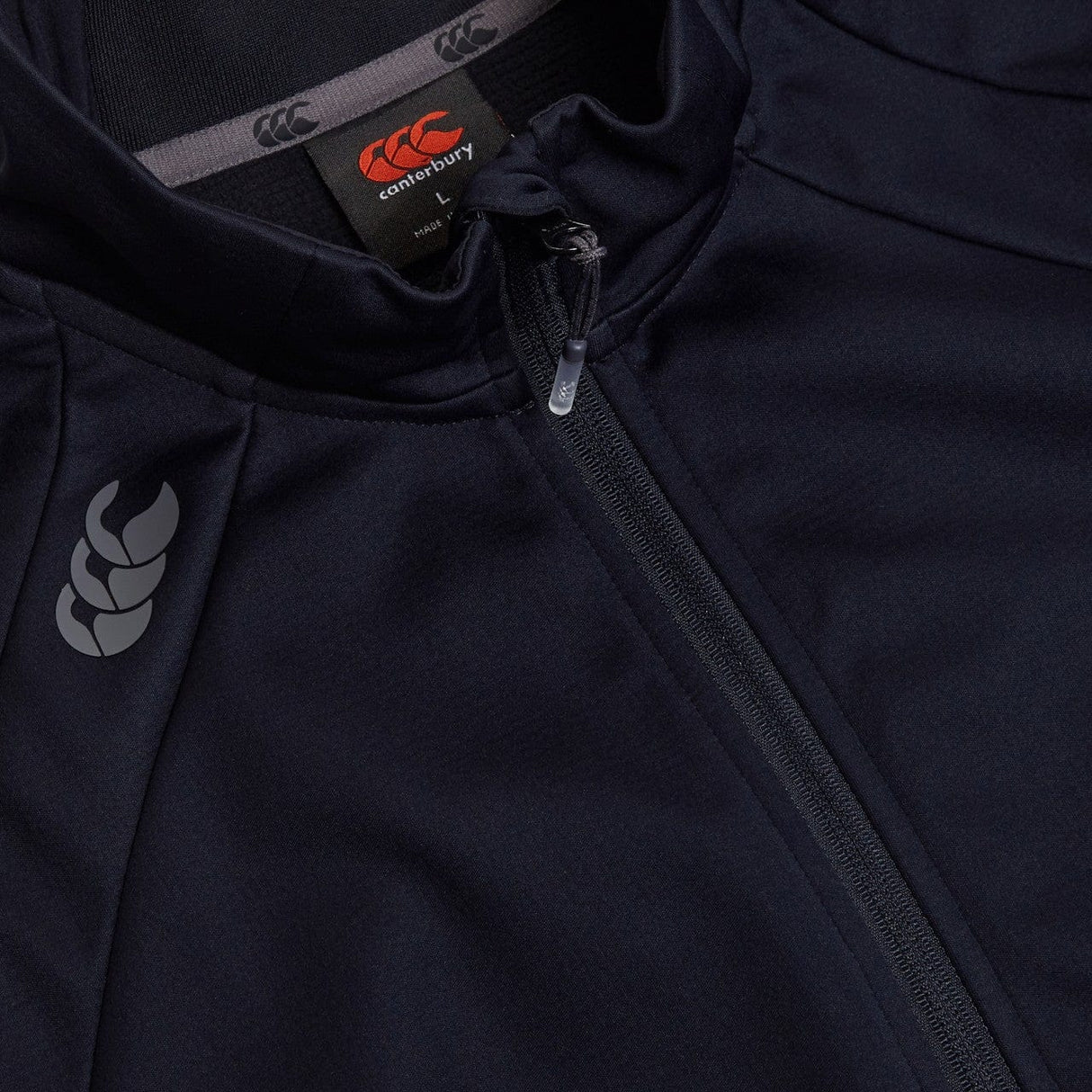 Close-up of a dark Canterbury Elite Windstopper Jacket with a Canterbury logo on the left chest and a partially unzipped front, designed for weatherproofing & mobility.