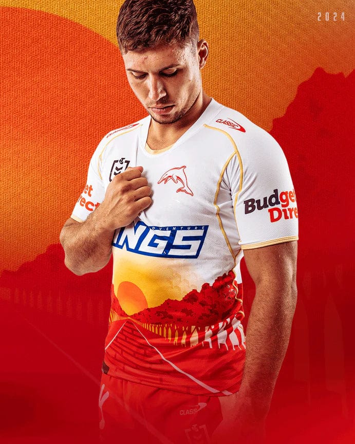 A man wearing a white and red sports jersey stands against a vibrant orange background, looking downward. The Dolphins NRL Heritage Jersey by Classic Sportswear features various logos, and the year 2024 is displayed in the top right corner.