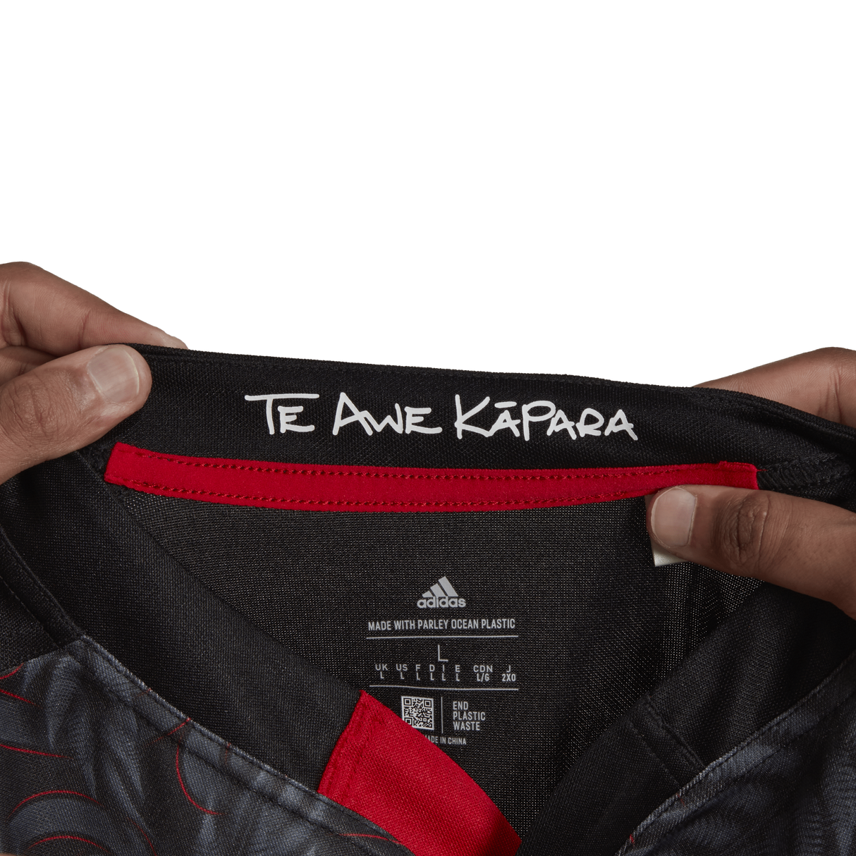 Hands holding the collar of a Māori All Blacks Home Supporters Jersey by adidas, featuring the text "Te Awe Kāpāra." The label showcases Adidas branding, washing instructions, and emphasizes its recycled polyester composition.