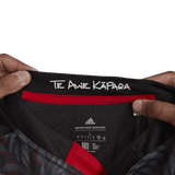 Hands holding the collar of a Māori All Blacks Home Supporters Jersey by adidas, featuring the text "Te Awe Kāpāra." The label showcases Adidas branding, washing instructions, and emphasizes its recycled polyester composition.