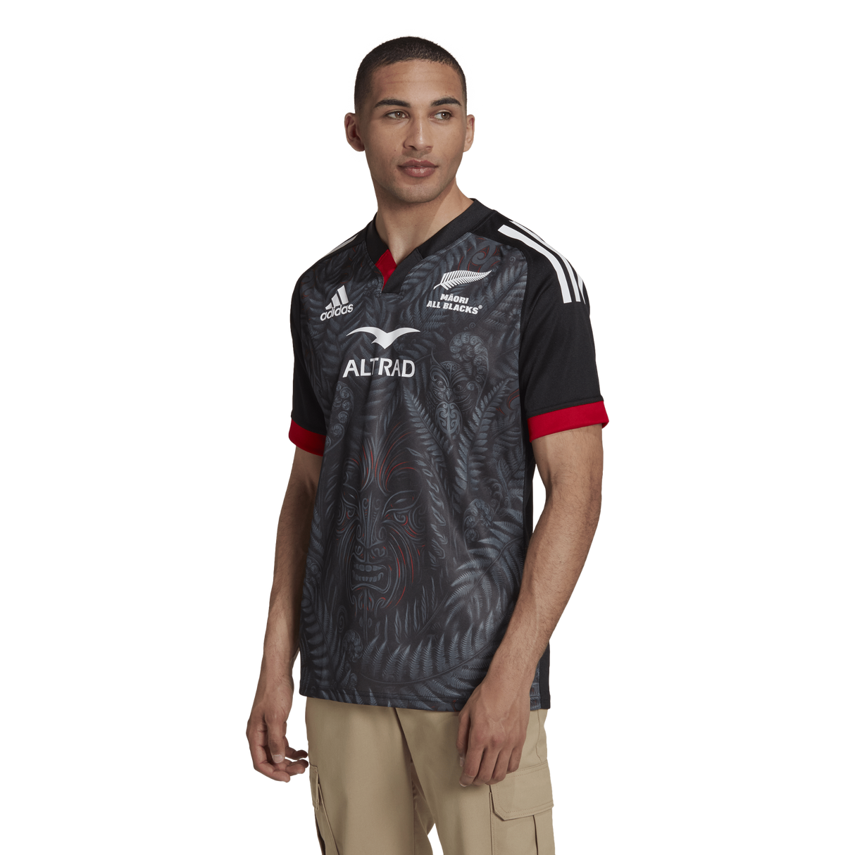 A man is wearing the Māori All Blacks Home Supporters Jersey by adidas, designed in black with red and white accents, featuring intricate patterns and logos, crafted from recycled polyester.