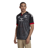 A man is wearing the Māori All Blacks Home Supporters Jersey by adidas, designed in black with red and white accents, featuring intricate patterns and logos, crafted from recycled polyester.