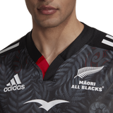 Wearing the adidas Māori All Blacks Home Supporters Jersey in black, a person proudly displays the white logos and intricate patterns on this moisture-wicking garment.