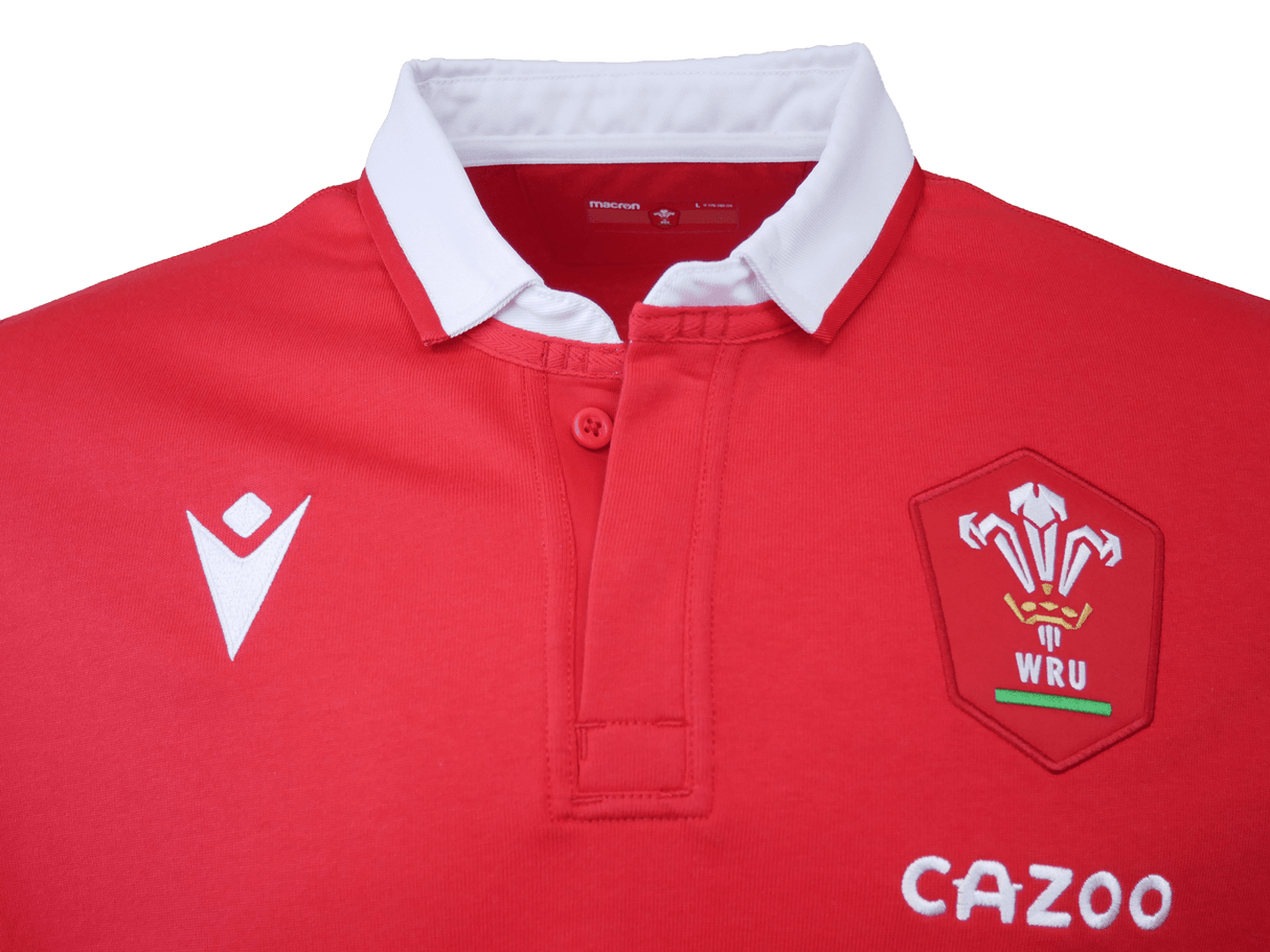 The Wales Long Sleeve Home Polo 22/23 by Macron is a red rugby jersey with a white collar, featuring the WRU logo, a white chevron emblem, and the sponsor logo "Cazoo." This classic rugby shirt is designed by Macron.