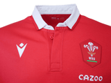 The Wales Long Sleeve Home Polo 22/23 by Macron is a red rugby jersey with a white collar, featuring the WRU logo, a white chevron emblem, and the sponsor logo "Cazoo." This classic rugby shirt is designed by Macron.